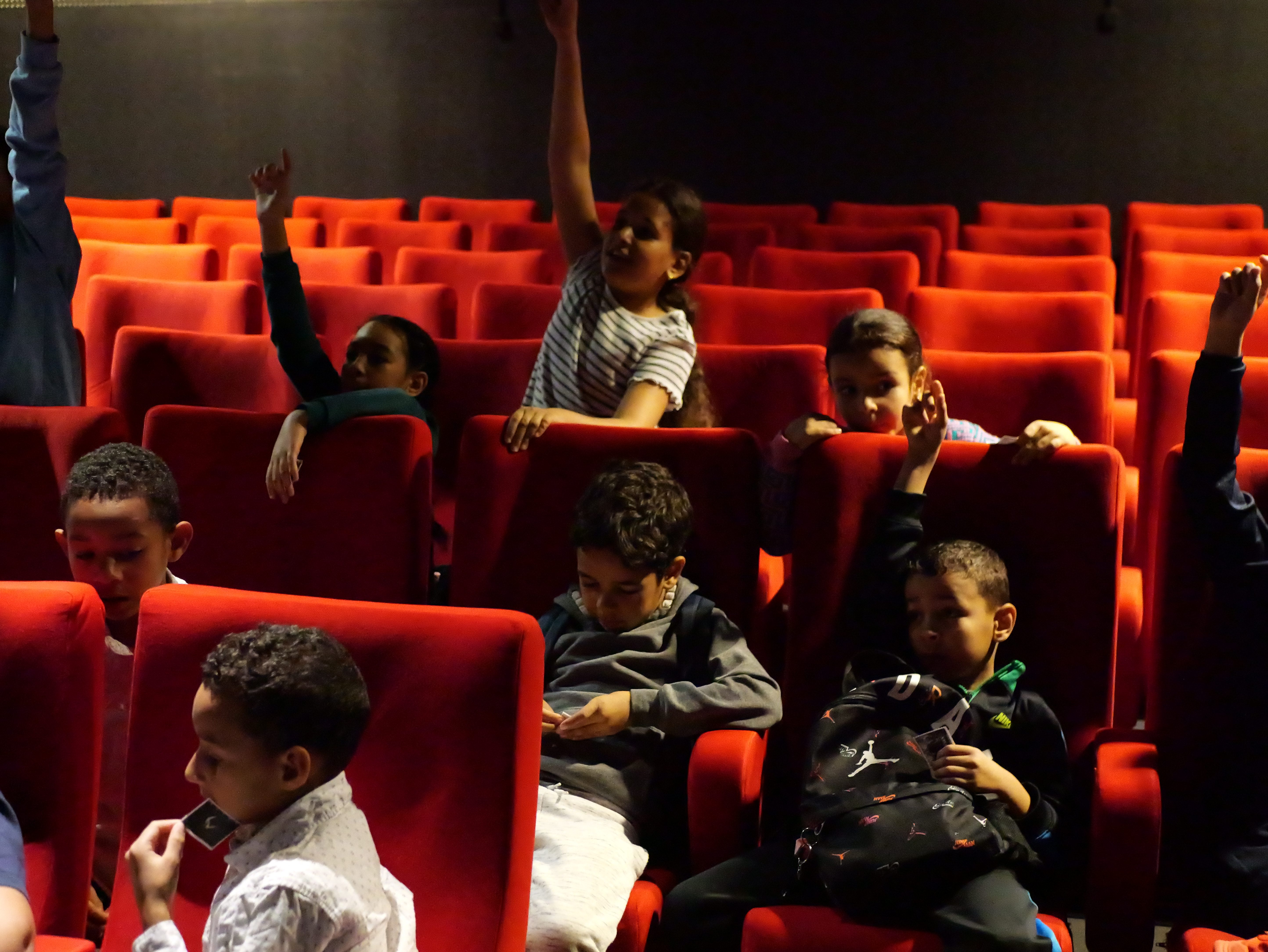 Les enfants du centre au cinéma, dans les fauteuils rouges, lèvent la main pour participer
